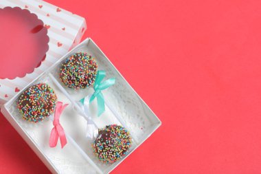 Renkli sprinkles ile çikolata Kek Pops. Kurdele yayı ile dekore edilmiştir. Şeffaf bir pencerenin kapağında bir hediye kutusunda yatıyorlar. Kırmızı arka planda.