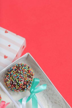 Renkli sprinkles ile çikolata Kek Pops. Kurdele yayı ile dekore edilmiştir. Şeffaf bir pencerenin kapağında bir hediye kutusunda yatıyorlar. Kırmızı arka planda.