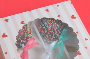 Renkli sprinkles ile çikolata Kek Pops. Kurdele yayı ile dekore edilmiştir. Şeffaf bir pencerenin kapağında bir hediye kutusunda yatıyorlar. Kırmızı arka planda.