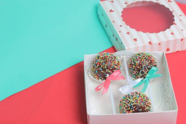 Renkli sprinkles ile çikolata Kek Pops. Kurdele yayı ile dekore edilmiştir. Şeffaf bir pencerenin kapağında bir hediye kutusunda yatıyorlar. Kırmızı ve naneli arka planda.