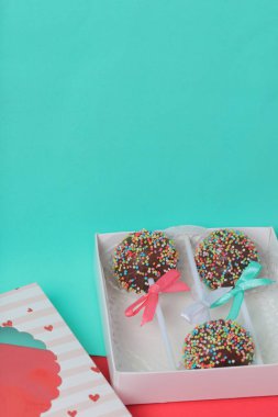 Renkli sprinkles ile çikolata Kek Pops. Kurdele yayı ile dekore edilmiştir. Şeffaf bir pencerenin kapağında bir hediye kutusunda yatıyorlar. Kırmızı ve naneli arka planda.