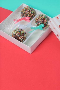 Renkli sprinkles ile çikolata Kek Pops. Kurdele yayı ile dekore edilmiştir. Şeffaf bir pencerenin kapağında bir hediye kutusunda yatıyorlar. Kırmızı ve naneli arka planda.