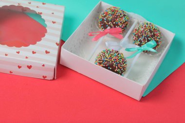 Renkli sprinkles ile çikolata Kek Pops. Kurdele yayı ile dekore edilmiştir. Şeffaf bir pencerenin kapağında bir hediye kutusunda yatıyorlar. Kırmızı ve naneli arka planda.