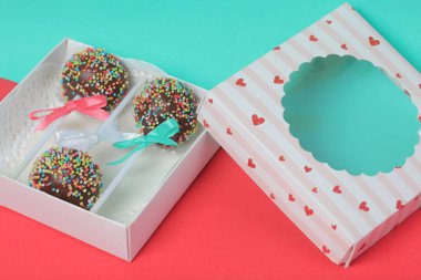 Renkli sprinkles ile çikolata Kek Pops. Kurdele yayı ile dekore edilmiştir. Şeffaf bir pencerenin kapağında bir hediye kutusunda yatıyorlar. Kırmızı ve naneli arka planda.