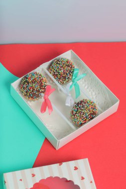 Renkli sprinkles ile çikolata Kek Pops. Kurdele yayı ile dekore edilmiştir. Şeffaf bir pencerenin kapağında bir hediye kutusunda yatıyorlar. Kırmızı ve naneli arka planda.