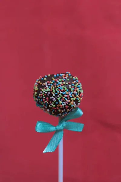 Bolo Pops em chocolate com polvilhas coloridas. Decorado com um laço de fita. Em um fundo vermelho — Fotografia de Stock