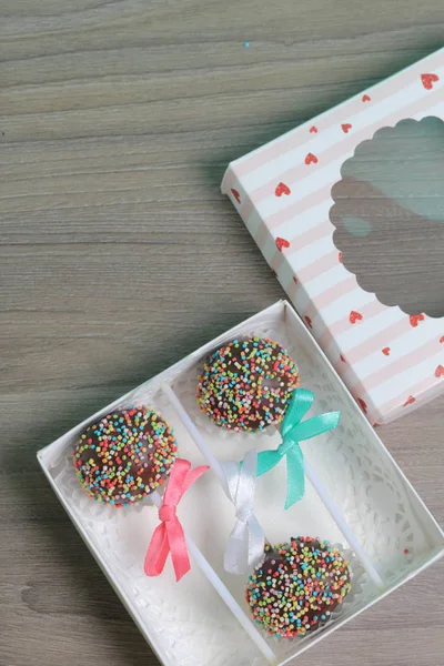 Cake Pops in chocolade met gekleurde hagelslag. Versierd met een strik van lint. Ze liggen in een geschenk doos, in het deksel waarvan er een doorzichtig venster is. — Stockfoto