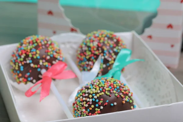 Cake Pops in chocolate with colored sprinkles. Decorated with a ribbon bow. They lie in a gift box, in the lid of which there is a transparent window.