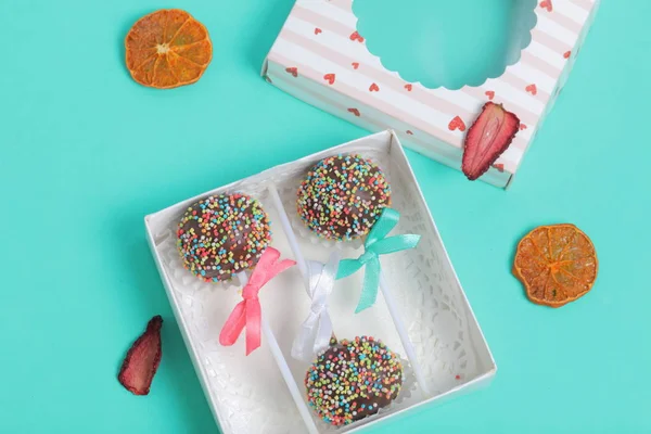 Pastel Pops en chocolate con chispas de colores. Decorado con un lazo de cinta. Se encuentran en una caja de regalo, en la tapa de la cual hay una ventana transparente. Muy cerca se encuentran los frutos secos para la decoración. En una menta — Foto de Stock