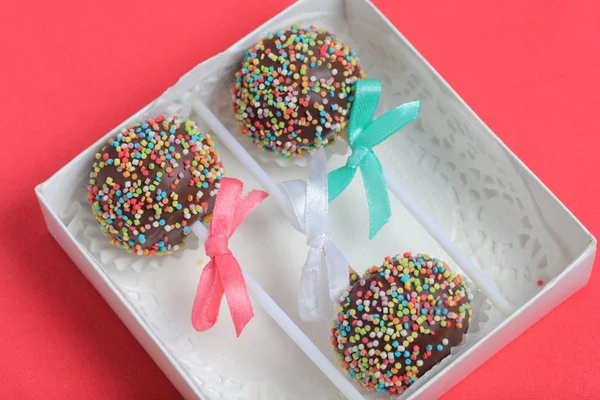 Cake Pops в шоколаде с цветными спреями. Украшенный ленточным бантом. Они лежат в подарочной коробке. На красном фоне . — стоковое фото