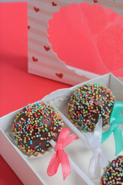 Pastel Pops en chocolate con chispas de colores. Decorado con un lazo de cinta. Se encuentran en una caja de regalo, en la tapa de la cual hay una ventana transparente. Sobre un fondo rojo . — Foto de Stock