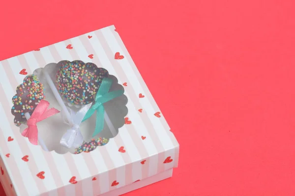 Pastel Pops en chocolate con chispas de colores. Decorado con un lazo de cinta. Se encuentran en una caja de regalo, en la tapa de la cual hay una ventana transparente. Sobre un fondo rojo . — Foto de Stock