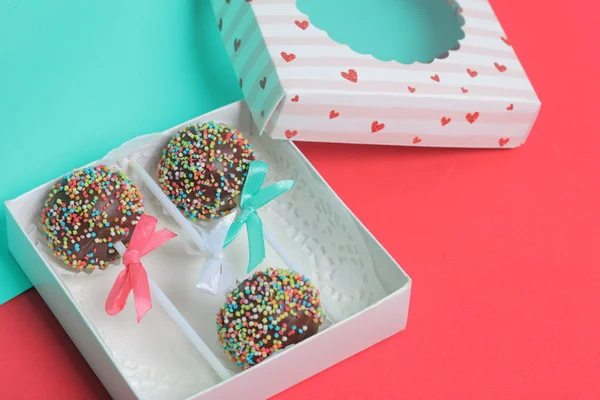 Pastel Pops en chocolate con chispas de colores. Decorado con un lazo de cinta. Se encuentran en una caja de regalo, en la tapa de la cual hay una ventana transparente. Sobre un fondo rojo y menta . — Foto de Stock