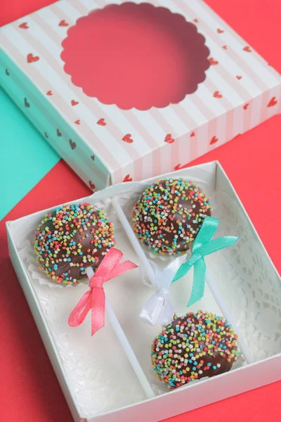 Cake Pops в шоколаде с цветными спреями. Украшенный ленточным бантом. Они лежат в подарочной коробке, в крышке которой есть прозрачное окно. На красном и мятном фоне . — стоковое фото