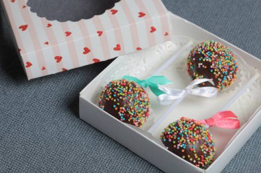 Renkli sprinkles ile çikolata Kek Pops. Kurdele yayı ile dekore edilmiştir. Şeffaf bir pencerenin kapağında bir hediye kutusunda yatıyorlar. Gri arka planda.