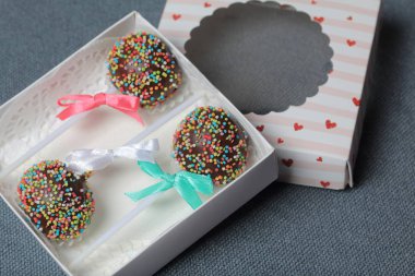 Renkli sprinkles ile çikolata Kek Pops. Kurdele yayı ile dekore edilmiştir. Şeffaf bir pencerenin kapağında bir hediye kutusunda yatıyorlar. Gri arka planda.