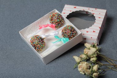 Renkli sprinkles ile çikolata Kek Pops. Kurdele yayı ile dekore edilmiştir. Şeffaf bir pencerenin kapağında bir hediye kutusunda yatıyorlar. Yakınlarda kurutulmuş güller vardır. Gri arka planda.