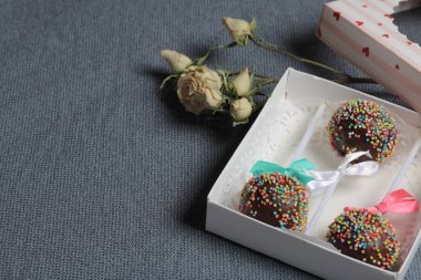 Renkli sprinkles ile çikolata Kek Pops. Kurdele yayı ile dekore edilmiştir. Şeffaf bir pencerenin kapağında bir hediye kutusunda yatıyorlar. Yakınlarda kurutulmuş güller vardır. Gri arka planda.