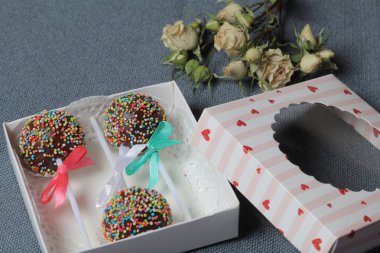 Renkli sprinkles ile çikolata Kek Pops. Kurdele yayı ile dekore edilmiştir. Şeffaf bir pencerenin kapağında bir hediye kutusunda yatıyorlar. Yakınlarda kurutulmuş güller vardır. Gri arka planda.
