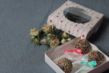 Renkli sprinkles ile çikolata Kek Pops. Kurdele yayı ile dekore edilmiştir. Şeffaf bir pencerenin kapağında bir hediye kutusunda yatıyorlar. Yakınlarda kurutulmuş güller vardır. Gri arka planda.