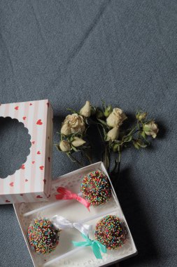 Renkli sprinkles ile çikolata Kek Pops. Kurdele yayı ile dekore edilmiştir. Şeffaf bir pencerenin kapağında bir hediye kutusunda yatıyorlar. Yakınlarda kurutulmuş güller vardır. Gri arka planda.