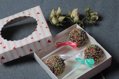 Renkli sprinkles ile çikolata Kek Pops. Kurdele yayı ile dekore edilmiştir. Şeffaf bir pencerenin kapağında bir hediye kutusunda yatıyorlar. Yakınlarda kurutulmuş güller vardır. Gri arka planda.