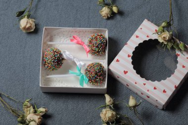 Renkli sprinkles ile çikolata Kek Pops. Kurdele yayı ile dekore edilmiştir. Şeffaf bir pencerenin kapağında bir hediye kutusunda yatıyorlar. Yakınlarda kurutulmuş güller vardır. Gri arka planda.