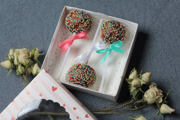 Pastel Pops en chocolate con chispas de colores. Decorado con un lazo de cinta. Se encuentran en una caja de regalo, en la tapa de la cual hay una ventana transparente. Cerca hay rosas secas. Sobre un fondo gris . — Foto de Stock