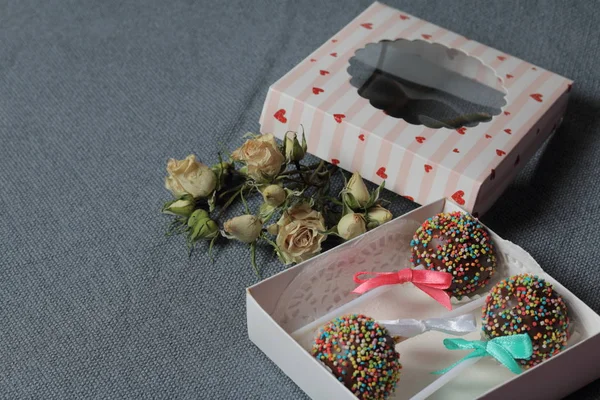 Cake Pops в шоколаде с цветными спреями. Украшенный ленточным бантом. Они лежат в подарочной коробке, в крышке которой есть прозрачное окно. Рядом высушенные розы. На сером фоне . — стоковое фото