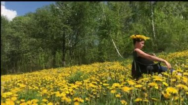 Bir kız piknik koltuğunda oturuyor. Bahar çayırında, çiçekli karahindibalarla büyümüş. Başında karahindiba çelengi var. Parkta güneşli bahar.