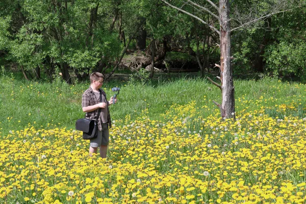 Jeune homme dodu dans une prairie avec des pissenlits en fleurs. Effectue l'enregistrement vidéo sur un smartphone installé sur un cardan. Près de la rivière coule . — Photo
