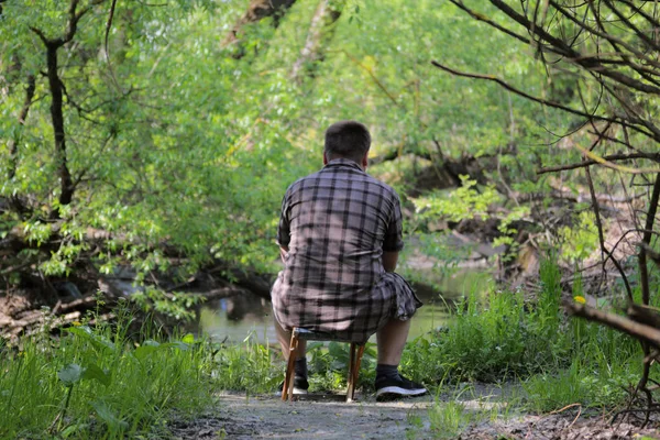 Um jovem gordo senta-se em uma pequena cadeira dobrável com as costas para a câmera. Sentado nas margens do rio a observar a água. No parque entre a folhagem verde brilhante . — Fotografia de Stock