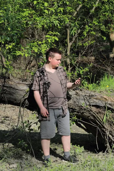 Un jeune homme dodu se tient près d'un arbre. Il tient un smartphone dans sa main. Pensivement en regardant le gadget . — Photo