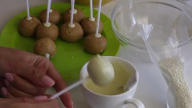 Bir kadın beyaz çikolata içine boş bir popcake kek dips ve topun yüzeyine eşit serpin. Sonraki masada popcakes yapmak için malzemeler vardır.