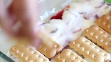 Bir kadın cam formda bir pasta üzerinde çilek dilimleri ekşi krema üstüne bisküvi bir tabaka döşeme olduğunu. Konteynerdeki kurabiyelerin yanında. Kurabiye, ekşi krema ve çilek kolay ve yumuşak kek.