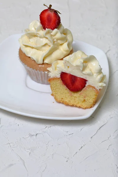 Cupcakes with strawberries and butter cream. One of them is cut, the filling is visible. On a white surface with decorative plaster. — Stock Photo, Image