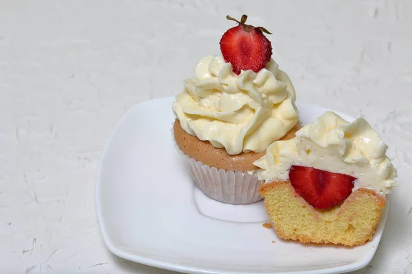 Cupcakes with strawberries and butter cream. One of them is cut, the filling is visible. On a white surface with decorative plaster. — Stock Photo, Image