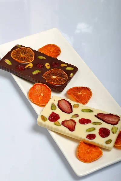 Homemade white and black chocolate. Decorated with slices of dried orange, strawberries, cherries and pistachios. On a white background in white ware.