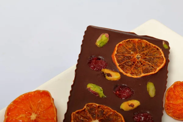 Homemade black chocolate. Decorated with slices of dried orange, strawberries, cherries and pistachios. On a white background in white ware.