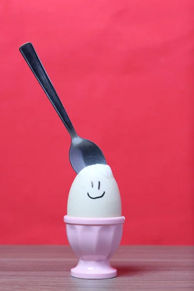 Ένα βραστό αυγό με κομμένο κέλυφος στέκεται σε ένα ποσέ. Στο αυγό που βάφτηκε χαμογελαστός. Ένα κουτάλι κόλλησε για φαγητό. Με φόντο το χρώμα των Κοραλλιών. — Φωτογραφία Αρχείου