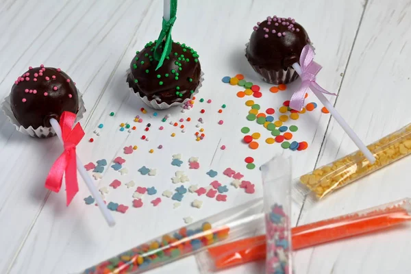 Cake pops decorated with a bow of braid. They lie on the boards, painted white. Nearby is scattered multicolored decorative topping. Visible topping in the package. — Stock Photo, Image