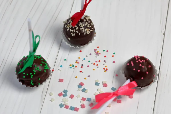 Cake pops decorated with a bow of braid. They lie on the boards, painted white. Nearby is scattered multicolored decorative topping. — Stock Photo, Image