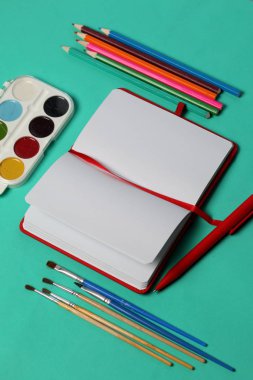 Kırmızı kapaktaki notlar için not defteri. Kırmızı bir yer imi ve kalemle. Nane arka planında açılan yalanlar. Sanat malzemeleri etrafında. Okul malzemeleri.