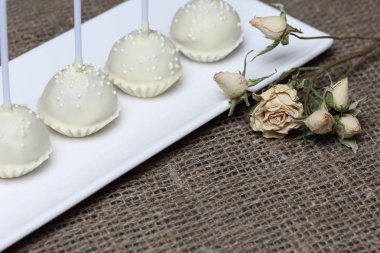 Kek beyaz çikolata sır açılır. Bir stand üzerinde dikey olarak durun. Beyaz dekoratif serpmelerle dekore edilmiştir. Kuru bej güle yakın. Kaba keten kumaş bir arka plan üzerinde.