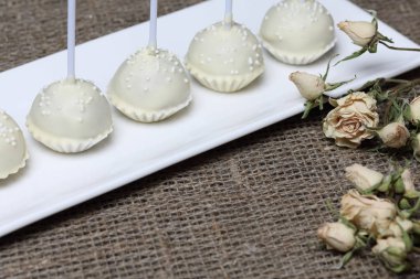 Kek beyaz çikolata sır açılır. Bir stand üzerinde dikey olarak durun. Beyaz dekoratif serpmelerle dekore edilmiştir. Kuru bej güle yakın. Kaba keten kumaş bir arka plan üzerinde.