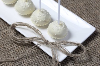 Kek beyaz çikolata sır açılır. Bir stand üzerinde dikey olarak durun. Beyaz dekoratif serpmelerle dekore edilmiştir. Sonraki yay, keten iplikten örülmüş. Kaba keten kumaş bir arka plan üzerinde.