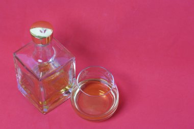Bir şişe güçlü alkol. Şeffaf camdan, altın stoperli kare bir form. Bir bardak yanında bir içki dökülür. Mercan renginin arka planına karşı.