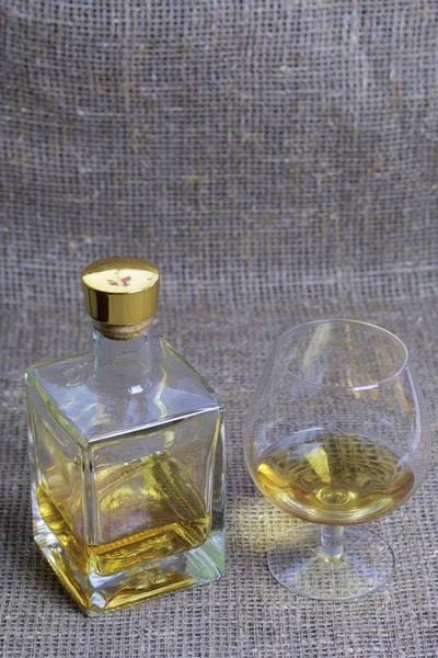 Una bottiglia di alcol forte. In vetro trasparente, forma quadrata con tappo dorato. Vicino a un bicchiere con una bevanda versata. Sullo sfondo di tessuto di lino grossolano . — Foto Stock