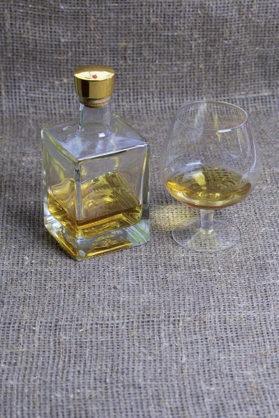 Бутылка крепкого алкоголя. Из прозрачного стекла, квадратной формы с золотой пробки. Возле стакана с налитой выпивкой. На фоне грубой льняной ткани . — стоковое фото