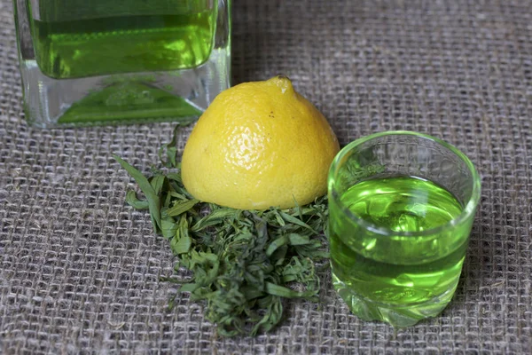 Silna Nalewka na Tarhuna w przezroczystej prostokątnej butelce. Pić jasny zielony wlewa się do szklanki. Obok składników do gotowania. Liści Estrada i cytryna. — Zdjęcie stockowe
